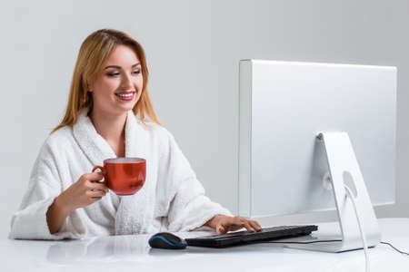 young woman sitting in the table and using computer on gray background. girl smiling and looking at monitor. with a cup in handsの写真素材