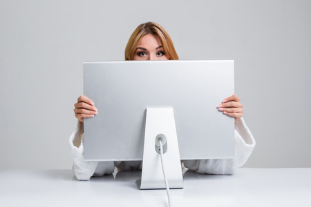 young woman sitting in the table and using computer on gray background. Surprised and scared hidden...