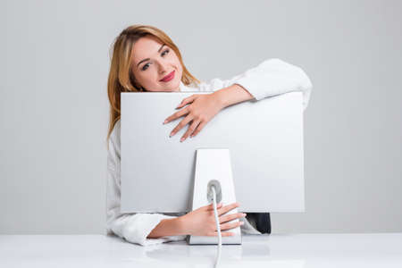 young woman sitting in the table and using computer on gray background. satisfied smiling girl hugging Monitorの写真素材