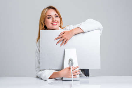 young woman sitting in the table and using computer on gray background. satisfied smiling girl hugging Monitorの写真素材