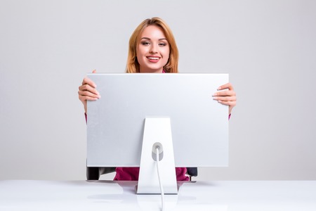 young woman sitting in the table and using computer on gray background. Surprised and scared hidden behind the monitor. It peeping from behind himの写真素材
