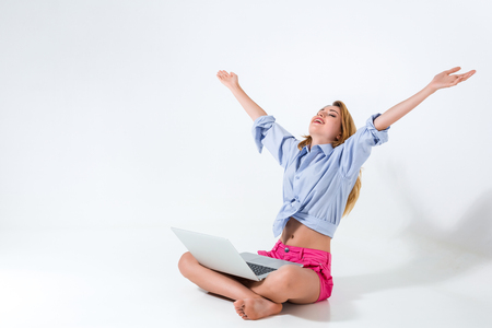 young woman sitting on the floor with crossed legs and using laptop on white background. very satisfied, happy, she raised her hands upの写真素材