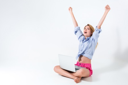 young woman sitting on the floor with crossed legs and using laptop on white background. very satisfied, happy, she raised her hands upの写真素材