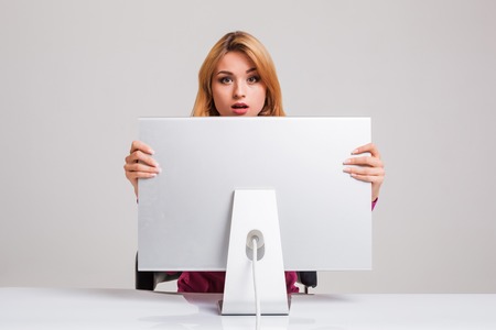 young woman sitting in the table and using computer on gray background. Surprised and scared hidden behind the monitor. It peeping from behind himの写真素材