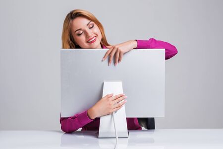 young woman sitting in the table and using computer on gray background. satisfied smiling girl hugging Monitorの写真素材