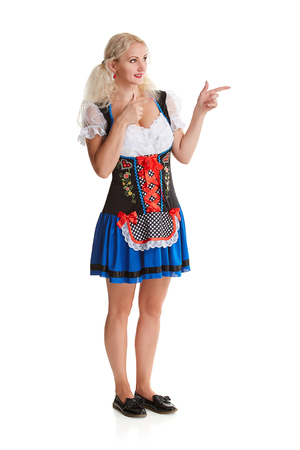 Beautiful young blond girl in dirndl drinks out of oktoberfest beer stein. Isolated on white background. It is a full-length. pointing finger and smiling. copyspace desing conceptの写真素材