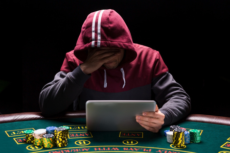 Online poker players sitting at the table. He play on tablet. The man in the hood, do not see the face. upset about losingの写真素材