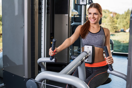 Muscular young brunette doing exercises on the simulator in the gym. She is wearing a gray tracksuitの写真素材