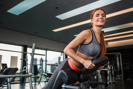 Muscular young brunette doing exercises on the simulator in the gym. She is wearing a gray tracksuit. Woman looking at the cameraの写真素材