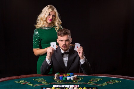 A young man in a suit shows a pair of aces. Playing poker at the table. Young woman enjoys winning at poker. They both look at the cameraの写真素材