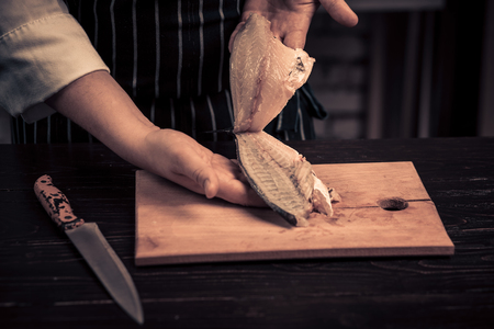 Chef cutting the fish on a board. Photo in brown tones. Kitchen. Wooden table. Hands of the cook close-upの写真素材