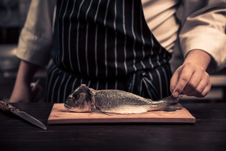 Chef cutting the fish on a board. Photo in brown tones. Kitchen. Wooden tableの写真素材