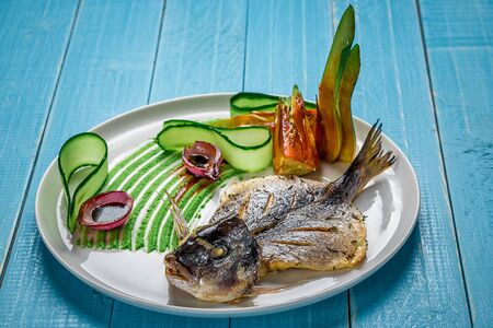 Fried fish dorado, decorated with cucumbers and onions. The original presentation of the dish from the chef. On a blue wooden backgroundの写真素材