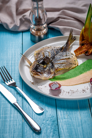 Fried fish dorado, decorated with cucumbers and onions. The original presentation of the dish from the chef. On a blue wooden backgroundの写真素材