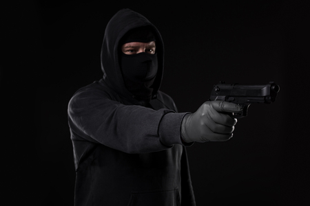 The robber in a mask with a gun pointed to the side on a black background. Man with a gunの写真素材