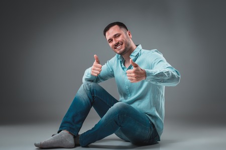 Handsome young man sitting on a floor with raised hands gesturing happiness on gray backgroundの写真素材
