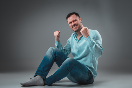 Handsome young man sitting on a floor with raised hands gesturing happiness on gray backgroundの写真素材