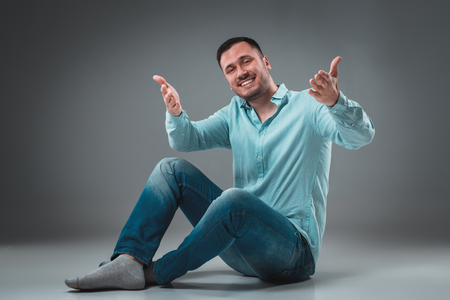 Handsome young man sitting on a floor with raised hands gesturing happiness on gray backgroundの写真素材