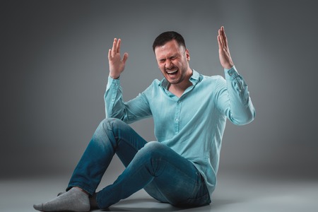 Handsome young man sitting on a floor with raised hands gesturing happiness on gray backgroundの写真素材