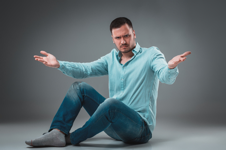 Handsome young man sitting on a floor with raised hands gesturing on gray backgroundの写真素材