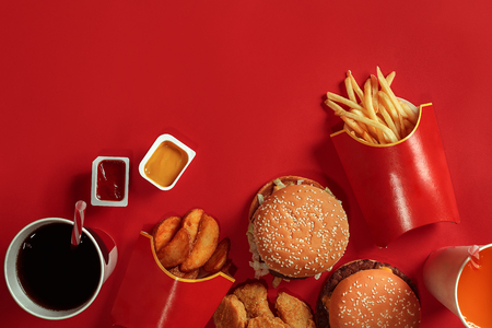 Two hamburgers and french fries, sauces and drinks on red background. Fast food. Top view, flat lay with copyspaceの写真素材