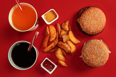 Two hamburgers and french fries, sauces and drinks on red background. Fast food. Top view, flat lay with copyspaceの写真素材