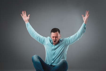 Casual young man sitting on the floor with his legs crossed and cheering with his hands in the air while screaming.の写真素材