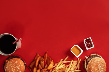 Fast food dish top view. Meat burger, potato chips and glass of drink on red background. Takeaway composition. Wrapped French fries, hamburger, mayonnaise and ketchup sauces on red desk. Copy spaceの写真素材