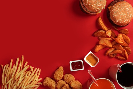 Fast food dish top view. Meat burger, potato chips and glass of drink on red background. Takeaway composition. Wrapped French fries, hamburger, mayonnaise and ketchup sauces on red desk. Copy spaceの写真素材
