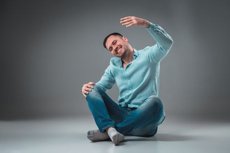 Handsome young man sitting on a floor with raised hands, isolated on gray backgroundの写真素材