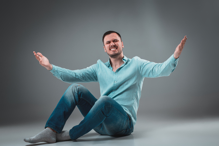 Handsome young man sitting on a floor with raised hands, isolated on gray backgroundの写真素材