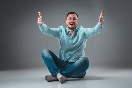 Handsome young man sitting on a floor with raised hands, isolated on gray backgroundの写真素材
