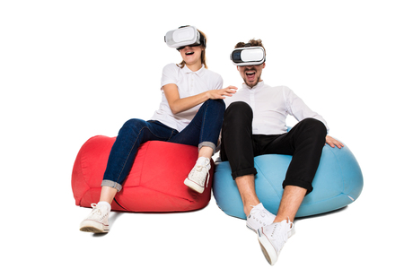 Excited young couple experiencing virtual reality seated on beanbags isolated on white background. A young couple dressed in jeans, white t-shirts and sneakersの写真素材