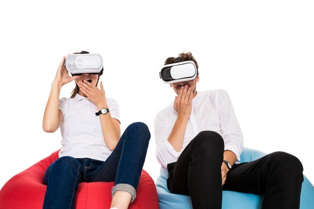 Excited young couple experiencing virtual reality seated on beanbags isolated on white background. A young couple dressed in jeans and white t-shirts, VRの写真素材