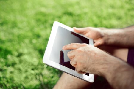 Close-up of casual dressed young man using modern digital tablet while sitting at the park, sun...