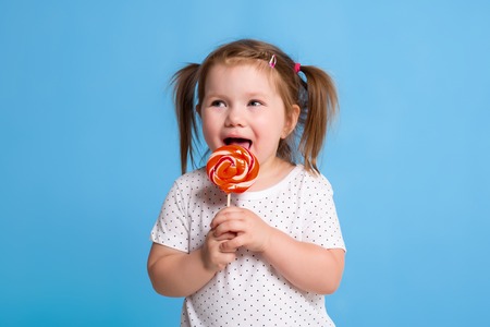 Beautiful little female child holding huge lollipop spiral candy smiling happy isolated on blue background.の写真素材