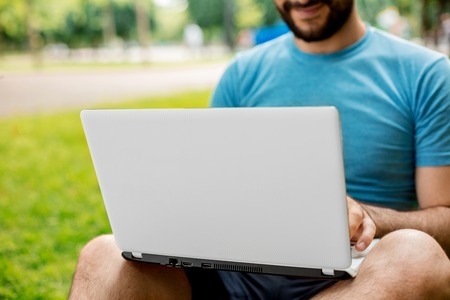 Young man using and typing laptop computer in summer grass. Freelancer working in outdoor parkの写真素材
