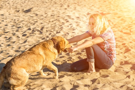 Portrait of young beautiful woman in sunglasses sitting on sand beach with golden retriever dog. Girl with dog by sea. Sun flareの写真素材