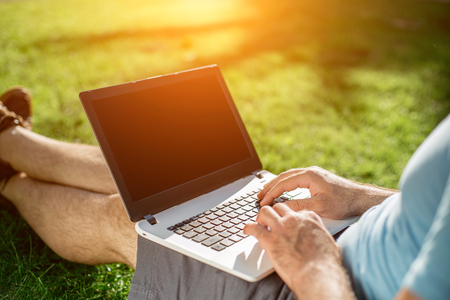 Cropped shot of man using laptop with blank screen while sitting on green grassの写真素材
