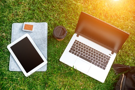 Laptop computer on green grass with coffee cup, bag and tablet in outdoor park