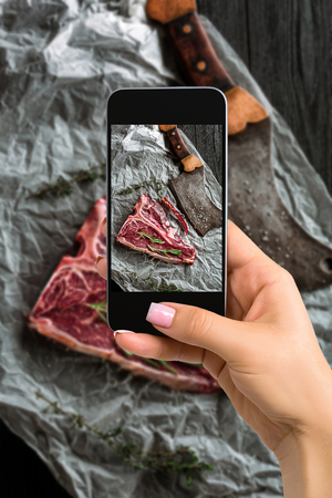 Photographing food concept - woman takes picture of raw dry aged t-bone steaks for grill with fresh herbs and cleaverの写真素材