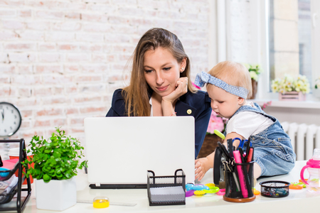 Cheerful young beautiful businesswoman looking at laptop while sitting at her working place with her little daughterの写真素材
