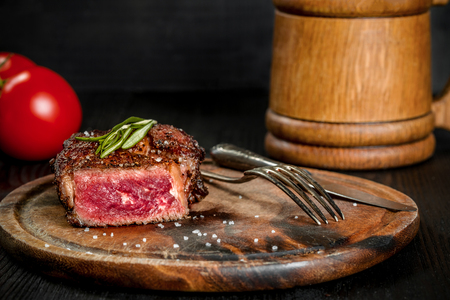 Grilled steak seasoned with spices and fresh herbs served on a wooden board with wooden mug of beer and fresh tomatoの写真素材