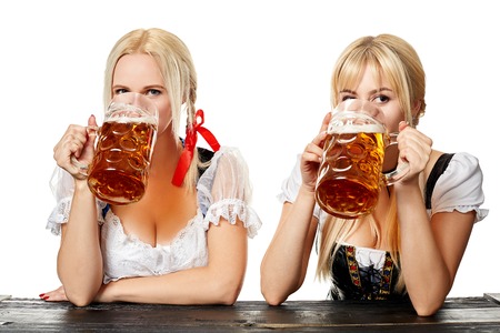 Two beautiful women drink beer from large glasses while sitting at a wooden table on a white background in the studioの写真素材