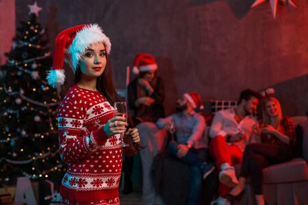 Portrait of a young woman with a glass of champagne at home on the foreground. Beautiful brunette in a Santa hat, red costume with deersの写真素材