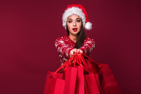 Young pretty woman or girl with long beautiful hair in Santa hat and New Years dress holding red package on red studio backgroundの写真素材