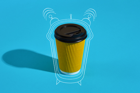 Hot coffee in yellow paper cup with black lid on blue background with shadow, blurred and soft focus image. Still life. Copy space. Wake up! Painted alarm clockの写真素材