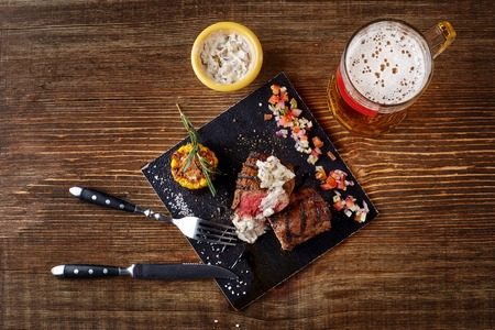 Grilled steak with corn with mushrooms sauce on cutting board and mug of beer on wooden table. Top viewの写真素材
