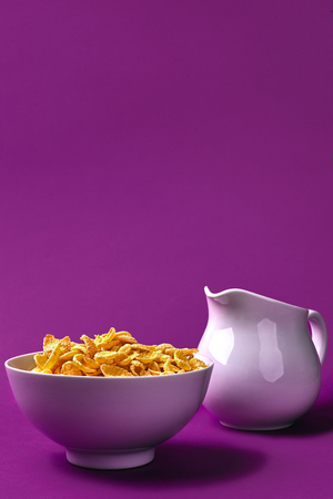 Bowl with corn flakes, jug of milk on purple background. oncept of a healthy breakfast. Copy space. Still life. Flat layの写真素材