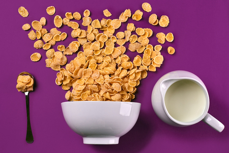 Bowl with corn flakes, jug of milk and spoon on purple background, top view
